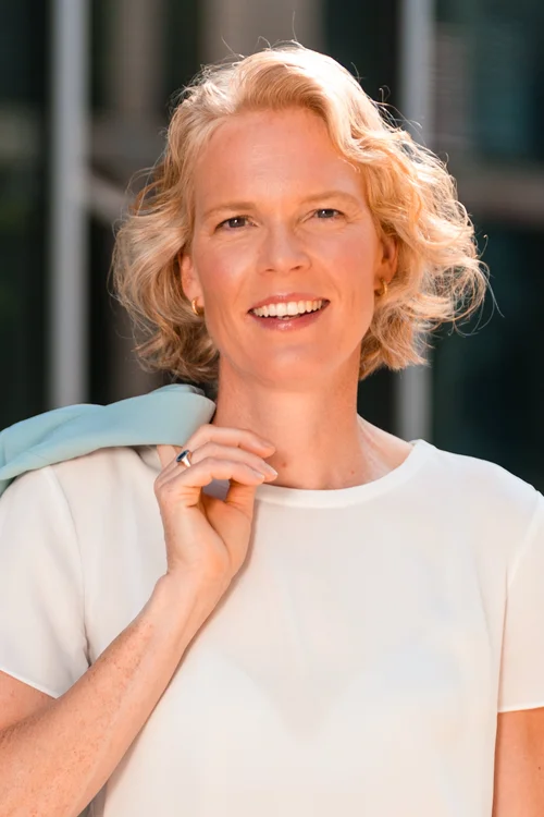 Porträt von Prof. Katharina Hölzle. Eine Frau gelesene Person mit blonden Haaren und einem weißen T-Shirt lächelt in die Kamera.