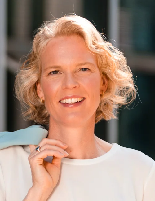 Porträt von Prof. Katharina Hölzle. Eine Frau gelesene Person mit blonden Haaren und einem weißen T-Shirt lächelt in die Kamera.