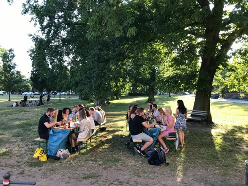 Zwei Bierbänke auf einer Wiese mit Bäumen, an denen Studierende sitzen, die essen und trinken.