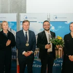 Vier Personen, eine hält eine Statuette und einen Blumenstrauß