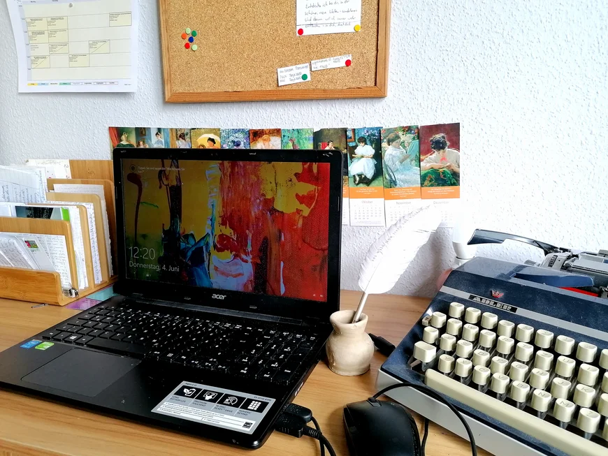 Auf einem Schreibtisch steht ein Laptio und daneben eine alte Schreibmaschine sowie eine Schreibfeder. An der Wand dahinter hängt eine Pinnwand und Kalenderblätter mit kleinen Gemälden