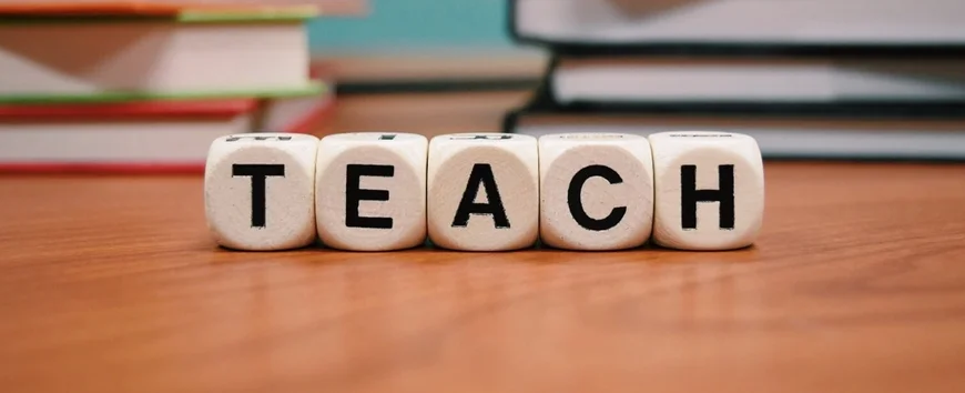 Fünf Würfel in einer Reihe mit jeweils einem Buchstaben, die das Wort Teach ergeben. Im Hintergrund sind Bücher zu sehen.