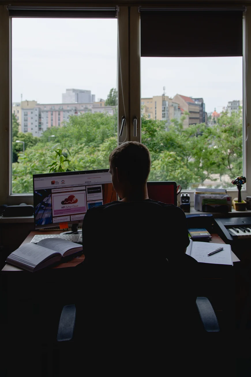 Ein Student sitzt an seinem Schreibtisch vor einem großen Fenster. Draußen sieht man Bäume und große Hochhäuse. Vor sich hat er ein Buch und seinen Computer