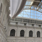 Ein Blick nach oben in das gläserne Dach des Archäologischen Museums durch welches blauer Himmel zu erkennen ist. Die Metall-Glaskonstruktion wird getragen von weißen Wänden, die rhythmisch auf mehreren Etagen durch Torbögen unterbrochen sind.