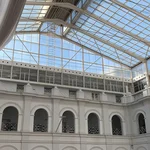 Ein Blick nach oben in das gläserne Dach des Archäologischen Museums durch welches blauer Himmel zu erkennen ist. Die Metall-Glaskonstruktion wird getragen von weißen Wänden, die rhythmisch auf mehreren Etagen durch Torbögen unterbrochen sind.