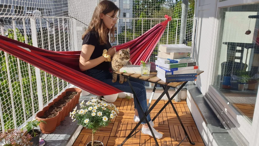 Eine junge Frau sitzt in einer roten Hängematte, die über den Balkon gespannt ist, auf dem Schoß eine flauschige Katze. Vor ihr auf einem Tisch liegt ein großer Bücherstapel, danben ein mit Minze dekoriertes Getränk