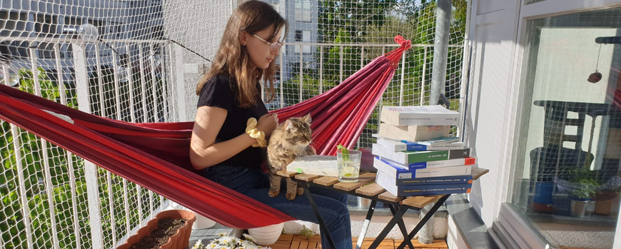 Eine junge Frau sitzt in einer roten Hängematte, die über den Balkon gespannt ist, auf dem Schoß eine flauschige Katze. Vor ihr auf einem Tisch liegt ein großer Bücherstapel, danben ein mit Minze dekoriertes Getränk