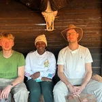 drei Menschen sitzen in einer Holzhütte unter einem Tierschädel