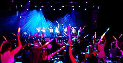 Partybeleuchtung im Waschhaus. Viele Menschen auf Trampolinen, die auf die Bühne schauen auf der ebenso Menschen auf Trampolinen sind. Leuchtstäbe werden in die Luft gehalten.