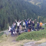 Gruppenfoto vor alpiner Bergkulisse. Die Menschen winken und geben Peace-Zeichen.