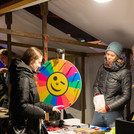 Drei Personen an einem Marktstand zwischen ihnen ein buntes Glücksrad