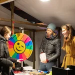 Drei Personen an einem Marktstand zwischen ihnen ein buntes Glücksrad