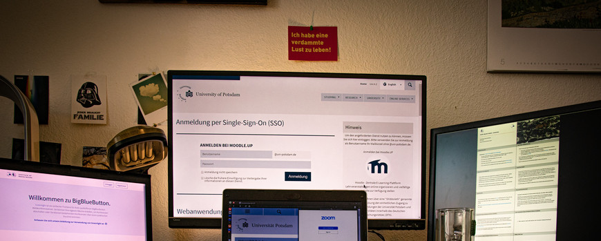 Auf einem Schreibtsch ein Laptop und drei Bildschirme. Auf einem ist Big Blue Button, ein Konferenzdienst, geöffnet. Auf dem anderen die Single sign On Seite der UP. Auf einem weiteren eine Sammlung Notizen. Auf dem Laptpop eine Präsentation mit dem Titel "Die Corona Krise: Deutungen Folgen Chancen"