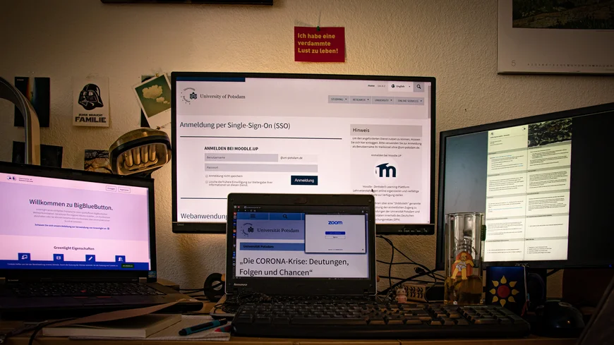 Auf einem Schreibtsch ein Laptop und drei Bildschirme. Auf einem ist Big Blue Button, ein Konferenzdienst, geöffnet. Auf dem anderen die Single sign On Seite der UP. Auf einem weiteren eine Sammlung Notizen. Auf dem Laptpop eine Präsentation mit dem Titel "Die Corona Krise: Deutungen Folgen Chancen"