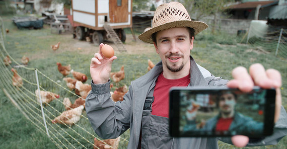 Landwirt mit Strohhut hält ein Ei in der einen und ein Smartphone in der anderen an und filmt sich für ein YouTube-Video.