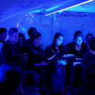 Zu sehen der Jazzchor Campus Golm in Aktion bei einem Konzert an der Universität Potsdam im StudentInnenkeller Nil 2023. Passend zum zum bläulich angestrahltem Gewölbe sind alle Sänger*innen dunkelblau gekleidet. Im Vordergrund sind die Besucher*innen zu sehen, die gespannt zuhören.