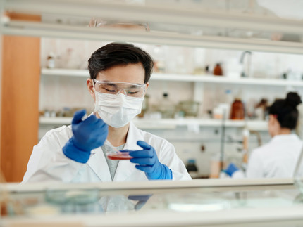 Eine Person mit Mundschutz und Schutzbrille arbeitet in einem Labor. Die Person hält eine Petrischale und eine Pipette in der Hand.