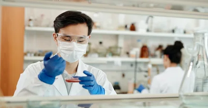 Eine Person mit Mundschutz und Schutzbrille arbeitet in einem Labor. Die Person hält eine Petrischale und eine Pipette in der Hand.
