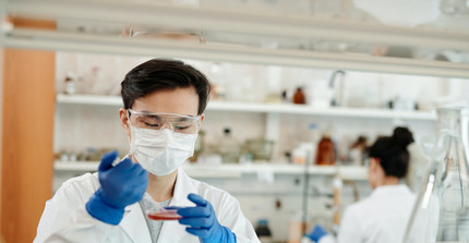Eine Person mit Mundschutz und Schutzbrille arbeitet in einem Labor. Die Person hält eine Petrischale und eine Pipette in der Hand. 