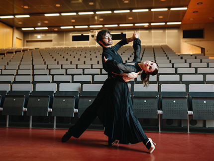 Zwei Personen in schwarzer Kleidung machen eine Tanzbewegung, im Hintergrund sieht man Stühle eines Hörsaals