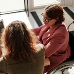 Das Bild ist in der Vogelperspektive, zwei Frauen sitzen an einem Tisch. Die einer Frau sitzt im Rollstuhl und trägt eine Brille. Von der anderen Frau sehen wir nur den Hinterkopf. Sie sitzen gemeinsam vor einem Laptop und arbeiten.