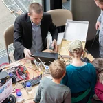 Eine Person sitzt mit Laptop an einem Tisch, mehrere Personen stehen davor.