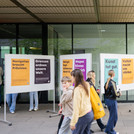 Mehrere Posterwände vor einem Gebäude mit Postern in orange, lila, hellblau und schwarz.