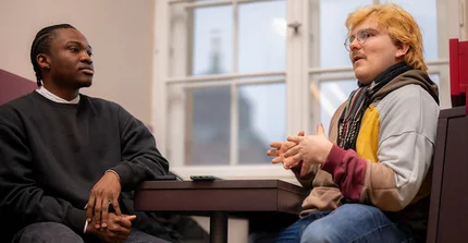 Two students are sitting casually across from each other at a table. One student is explaining something, using his hands, while the other student is listening.