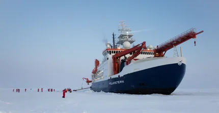Das Forschungsschiff "Polarstern" im ewigen Eis. Davor auf der Eisfläche sehr klein wirkend Menschen in grellroten Schneeanzügen.