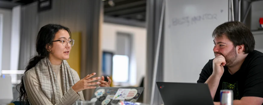 Eine Frau und ein Mann sitzen gemeinsam am Tisch und unterhalten sich gestikulierend. Im Hintergrund ist ein Lehrraum mit Whiteboard zu sehen.