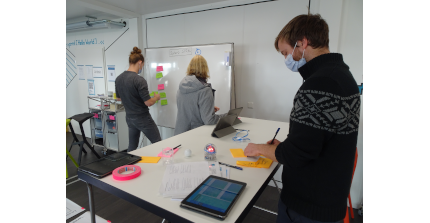 Drei Personen notieren in einem Workshopraum ihre Gedanken auf Zetteln und pinnen diese an ein Whiteboard.