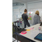Drei Personen notieren in einem Workshopraum ihre Gedanken auf Zetteln und pinnen diese an ein Whiteboard.