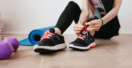 Person in Sportkleidung bindet sich den Shcuh zu. Neben ihr liegen Hanteln und eine Yogamatte.