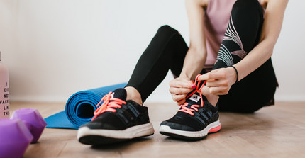 Person in Sportkleidung bindet sich den Shcuh zu. Neben ihr liegen Hanteln und eine Yogamatte. 