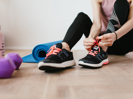 Person in Sportkleidung bindet sich den Shcuh zu. Neben ihr liegen Hanteln und eine Yogamatte.