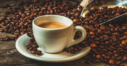 Eine weiße kleine Kaffeetasse auf Unterteller steht im Fokus. Darum herum viele lose Kaffeebohnen.