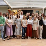 Ein Gruppenbild weißer Menschen in sommerlicher Kleidung, die freundlich in die Kamera schauen. IM Hintergrund ragt eine Skulptur des Berliner Bärs hervor.