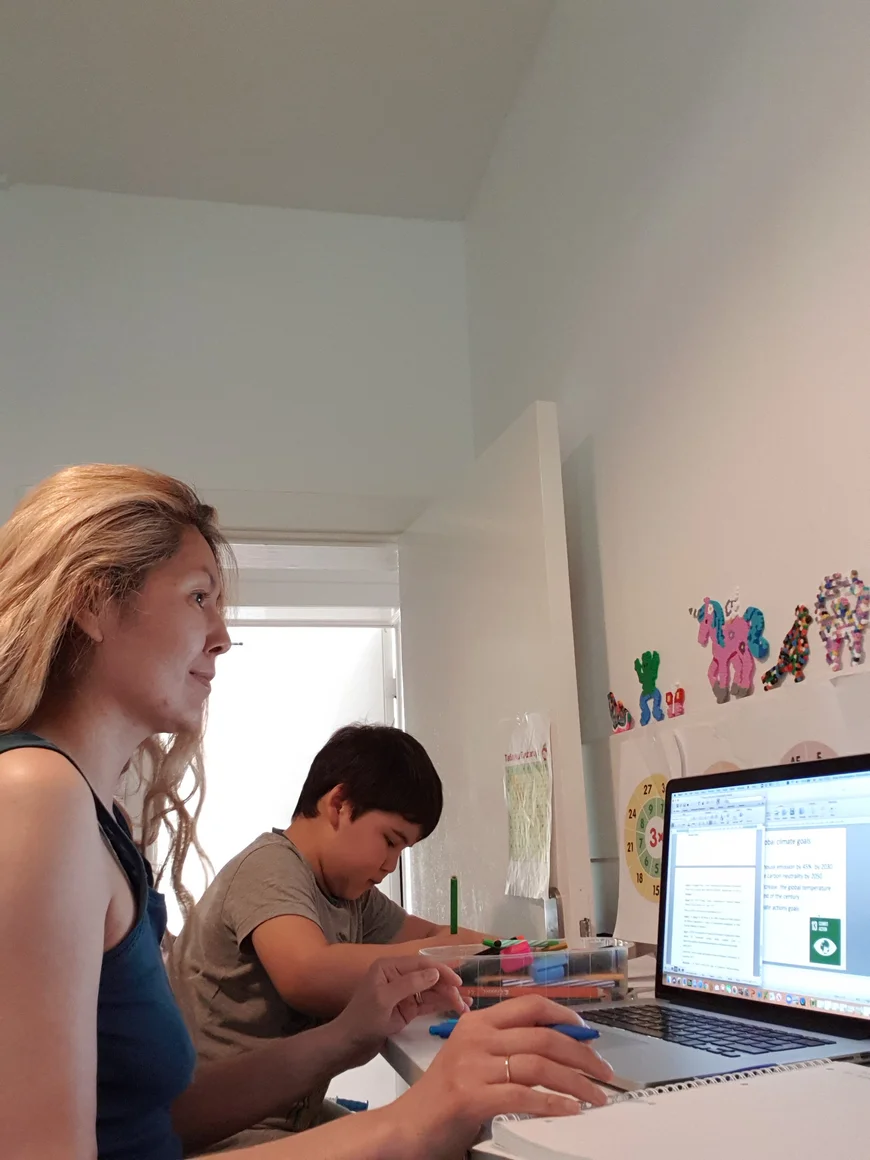 Eine Frau sitzt an einem Laptop und schreibt an einem Word Dokument. Neben ihr sitz ein ungefähr 10jähriger Junge der kontzentriert etwas malt oder schreibt.