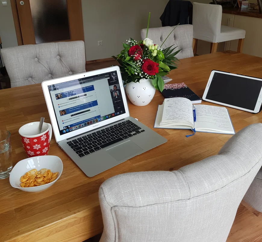 Auf einem Esstisch mit 4 Stühlen dran steht ein Mac book, ein Blumenstraß, eine Schüssel mit Crackern, eine rote Kaffeetasse. Daneben liegen Notizbücher und ein Tablet