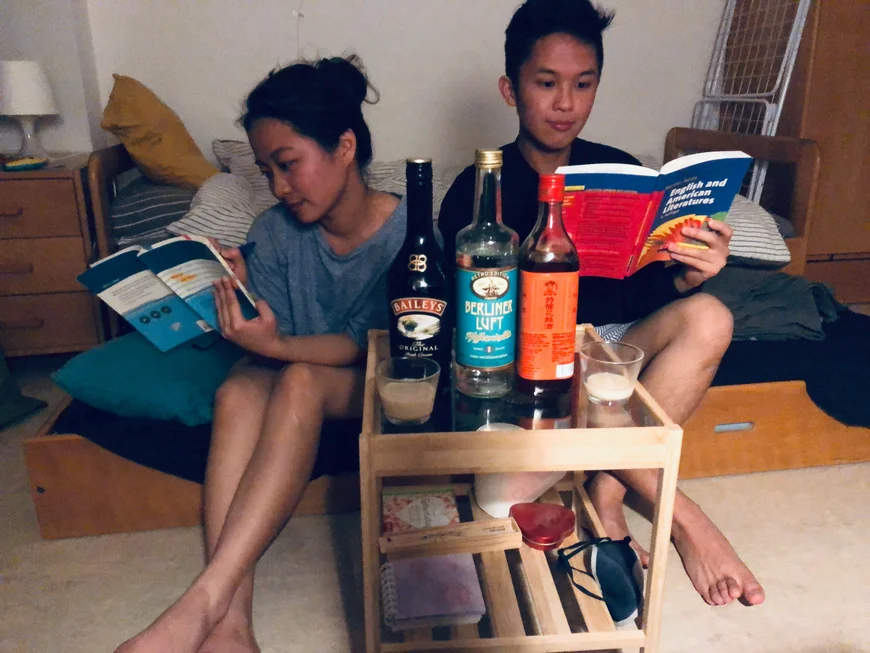 Zwei Studis hocken auf einem Bett und lesen jeweils ein Fachbuch. Vor ihnen steht ein Tischchen, darauf zwei gefüllte Gläser, eine Flasche Baylies, eine Flasche Berliner Luft und eine Flasche Wein. Vermute ich.