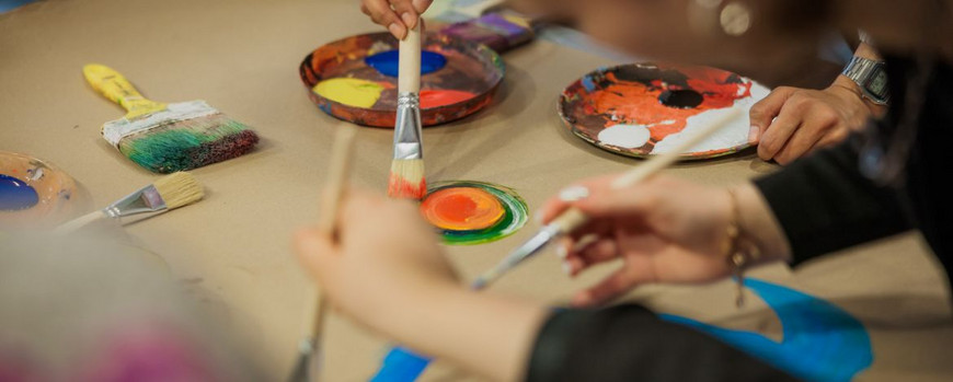 Hände mit Pinseln in der Hand. Gemiensam malen sie auf braunem Papier.