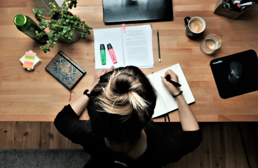 Ansicht von oben auf einen Schrebitisch, daran sietzt ein Menschen. Man sieht Kopf und Schultern, die Person schreibt in ein Notizbuch. Auf dem Tische Kaffeetasse, Wasserglas,Mouse, Stiftbecher, Textausdrucke, Notizbuch, Topfpflanze