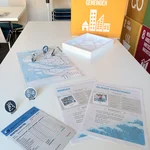 Das Bild zeigt das Relief des Deltas mit der entsprechenden Abbildung des Spielplans. Auf dem Spielplan sind Spielfiguren aufgestellt, die Sirenen, Mauern und Fluchtrouten darstellen und Teil des Spiels sind. Im Vordergrund sind Informationen zu vulnerablen Gruppen und eine Bestellliste zu sehen.