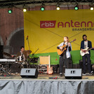 Sängerin mit Gitarre auf einer Bühne, im Hintergrund Gitarrist und Schlagzeuger