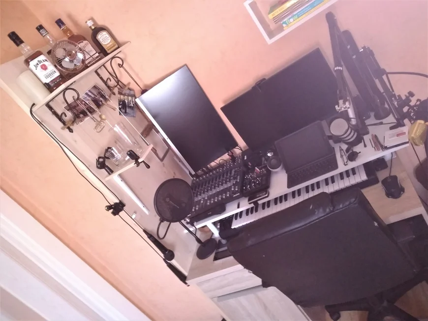 Ein Schreibtisch, darauf stehen sehr eng aneinander: Ein Laptop, zwei Bildschirme, ein Mischpult, zwei Mikrophone, ein Synthesizer, auf dem Regal links daneben ist enie Sammlung halbwegs trinkbarer Whiskey platziert