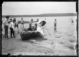 schwarz-weiß Foto einer Fotografin, die auf dem Rand eines Ruderboots sitzt und mit einer alten Kamera ein Foto machen will, dabei ist sie umringt von vielen Jungs, die teilweise in Badehosen im Wasser stehen oder bekleidet am Strand