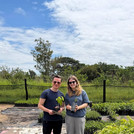 Auf dem Foto sind ein weißer junger Mann und eine weiße junge Frau im Freien zu sehen, die lächelnd zwischem grünen Baumsetzlingen stehen und zwei davon in die den Händen halten.