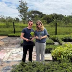 Auf dem Foto sind ein weißer junger Mann und eine weiße junge Frau im Freien zu sehen, die lächelnd zwischem grünen Baumsetzlingen stehen und zwei davon in die den Händen halten.