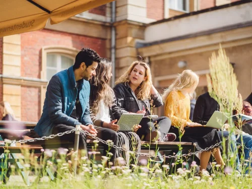 Studierende sitzen auf einer Bank am Campus Neues Palais und unterhalten sich. Eine Studentin hat einen Laptop auf dem Schoß.