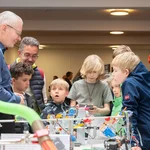 Mehrere Kinder und drei Erwachsene an einem Tisch auf dem mehrere technische Geräte stehen.